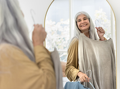 Older woman smiling while holding clothes in mirror, representing confidence and improved smile from cosmetic dentistry